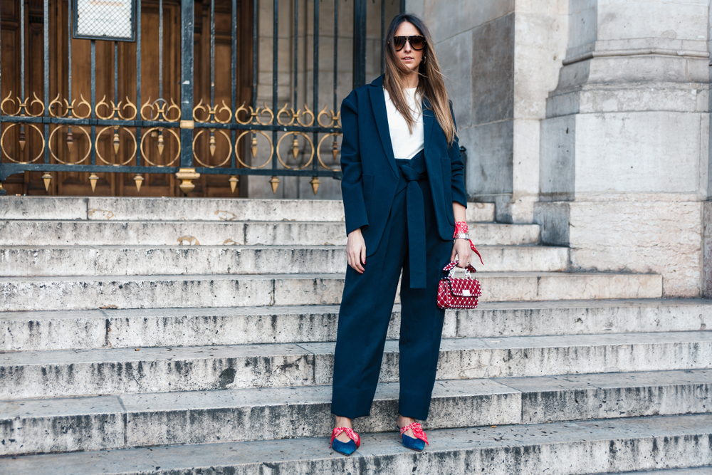 A woman in a blazer and matching paints with a white t-shirt stands on stairs in Paris, knowing what to wear to Paris in September, as she has a statement clutch as well.
