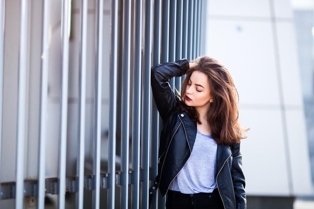 A woman in a gray shirt, leather jacket and jeans leans against a fence, knowing what to wear to Paris in September.