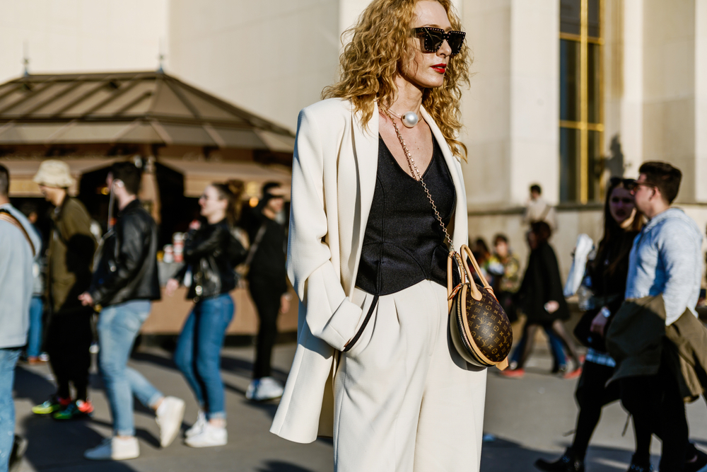 A woman wears a matching blazer and pants over a satin top with a giant pearl necklace and statement clutch.