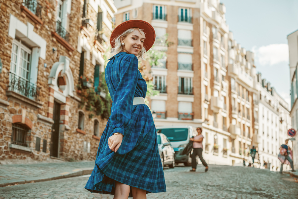 A girl in a plaid dress, belt and red hat walks down the streets of Paris and looks over her shoulder.