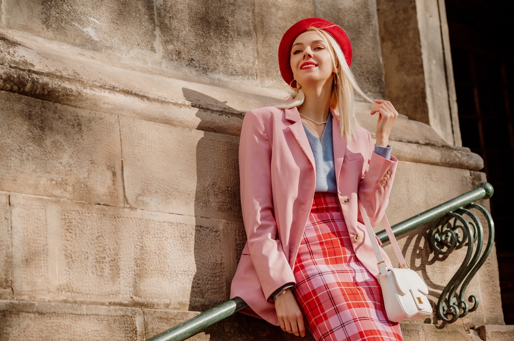 woman wearing stylish red beret, classic pink suit blazer, checkered skirt, holding trendy white bag, posing in street of European city. Article is about a 10 days in Europe packing list.