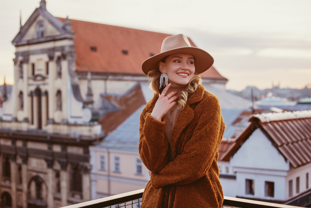 smiling woman wearing trendy beige hat, rhinestones earrings, brown faux fur coat, posing on balcony with beautiful view on European city.