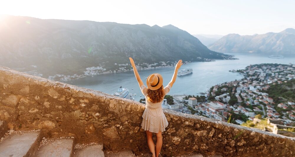 woman standing with her arms raised on stairs with stones along the railing overlooking a view of water in the background and towns lower down