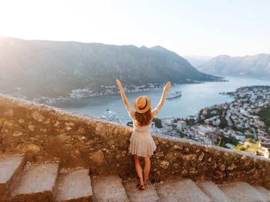 woman standing with her arms raised on stairs with stones along the railing overlooking a view of water in the background and towns lower down