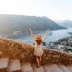 woman standing with her arms raised on stairs with stones along the railing overlooking a view of water in the background and towns lower down