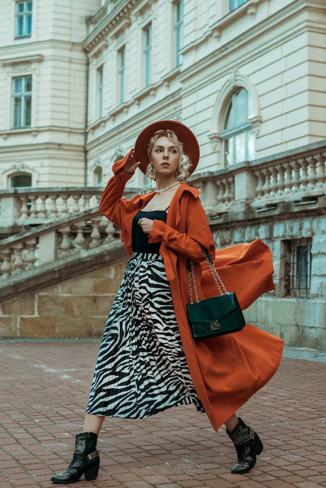Street style full-length photo of elegant woman wearing long orange trench coat, black silk top, hat, animal print midi skirt, cowboy ankle boots, with green bag, walking in street of European city