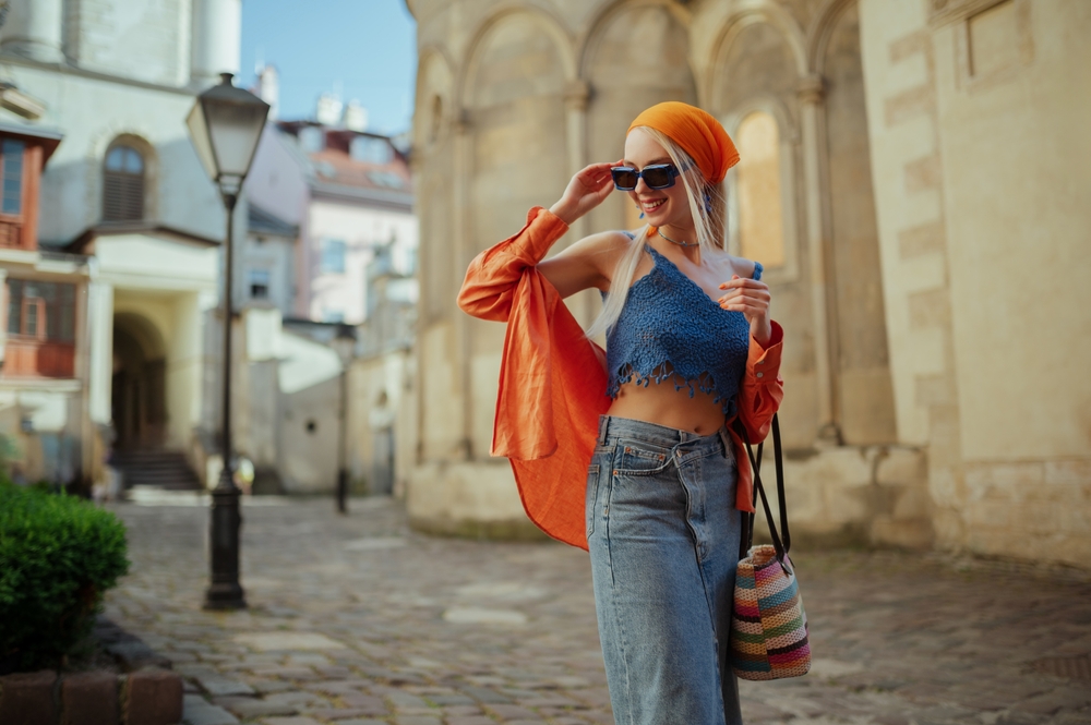 woman wearing trendy sunglasses, orange shirt, bandana, top, denim skirt, with wicker bag, posing in street of European city. Article is about a 10 days in Europe packing list.