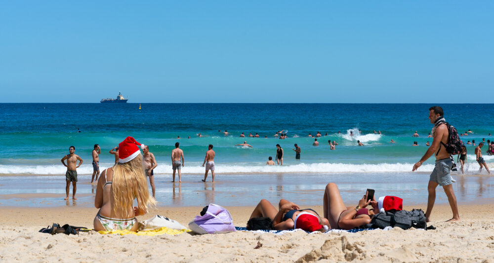 people wearing christmas hats and swimsuits sitting on the beach looking out at the ocean. the beach is sandy with tan sand and the ocean is blue with a blue sky