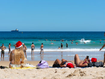 people wearing christmas hats and swimsuits sitting on the beach looking out at the ocean. the beach is sandy with tan sand and the ocean is blue with a blue sky