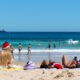 people wearing christmas hats and swimsuits sitting on the beach looking out at the ocean. the beach is sandy with tan sand and the ocean is blue with a blue sky