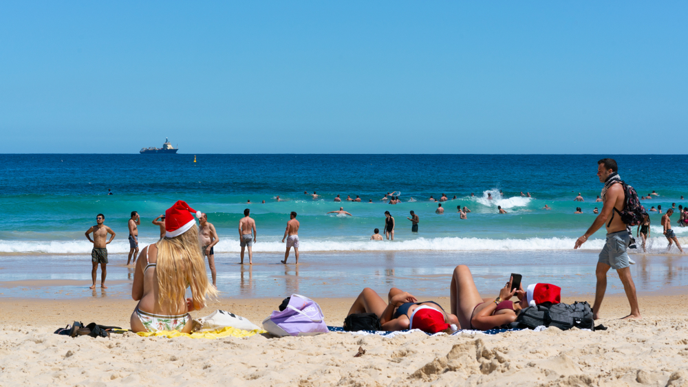 people wearing christmas hats and swimsuits sitting on the beach looking out at the ocean. the beach is sandy with tan sand and the ocean is blue with a blue sky