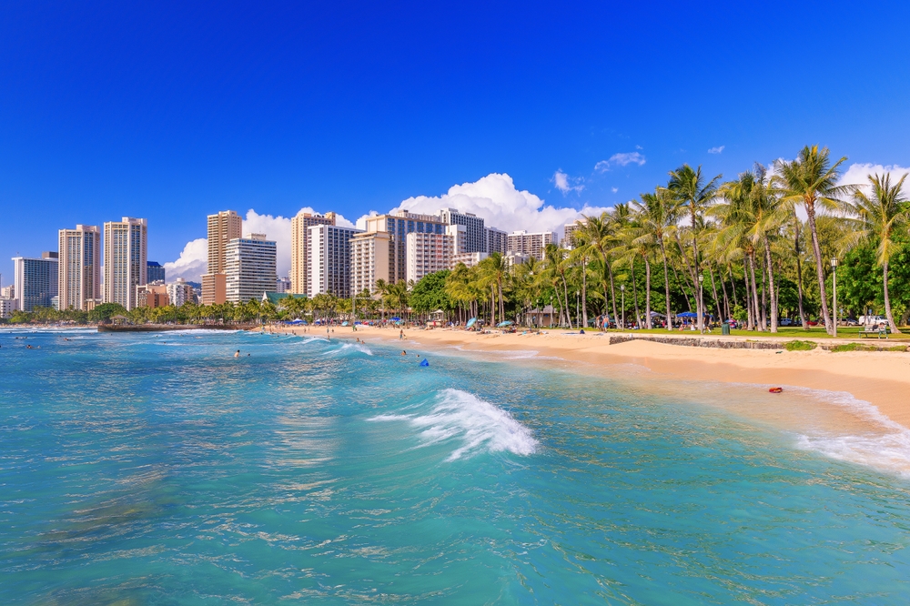 A shot of Honolulu shows crystal clear waters, warm sand, towering hotels, and green palm trees, perfect for relaxing and of the Best Places to Celebrate Christmas on The Beach.