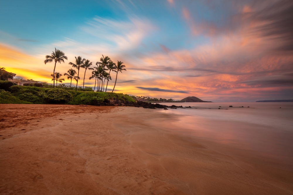 In Maui, a sunset hits the coast, palm trees tall, sand warm and water calm: this is one of the Best Places to Celebrate Christmas on The Beach.