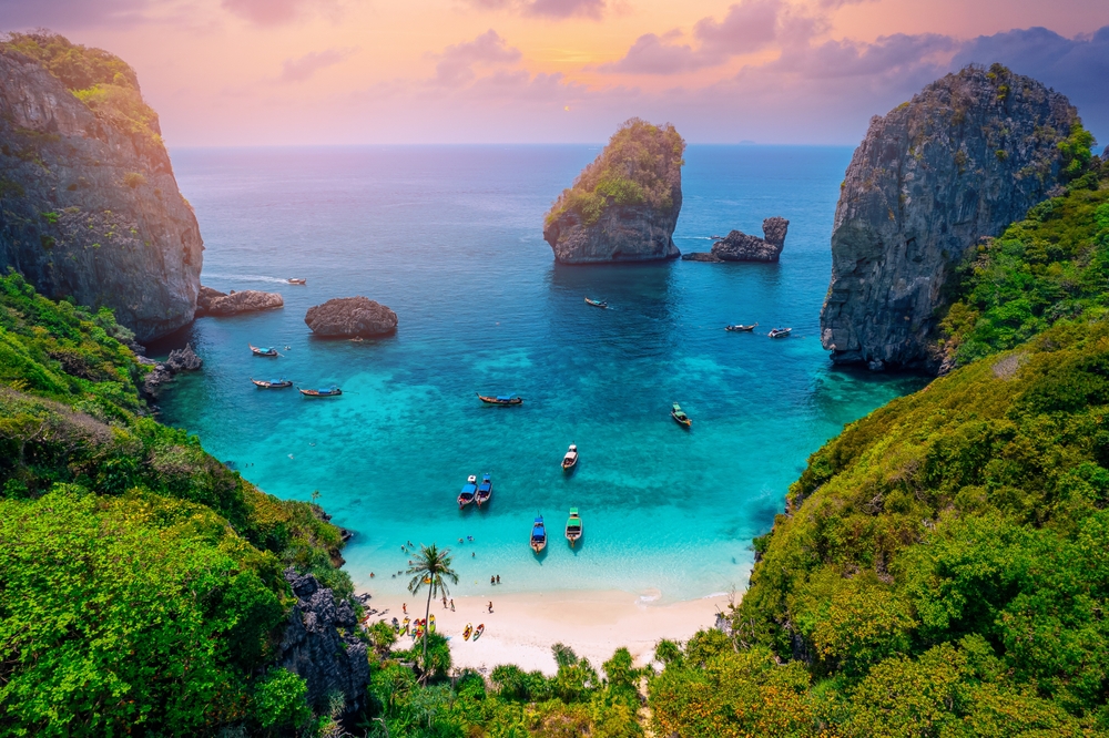 An overhead photo of Phuket shows a viewpoint from down green mountains and into a cove with white sand, blue waters, and docked boats.