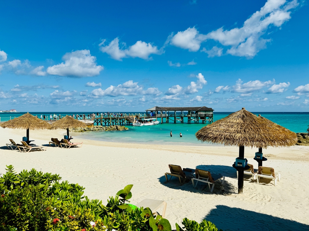 The Bahamas is well know for its powered sand beaches, and bright waters: in this photo, guests line a dock and tiki huts that feature chairs/loungers on the beach.
