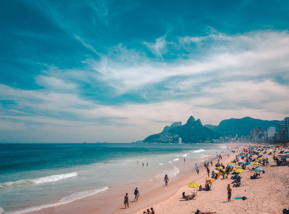 In Rio De Janerio, tourists line the beach with blue and green umbrellas, mountains in the distance: this is one of the Best Places to Celebrate Christmas on The Beach.