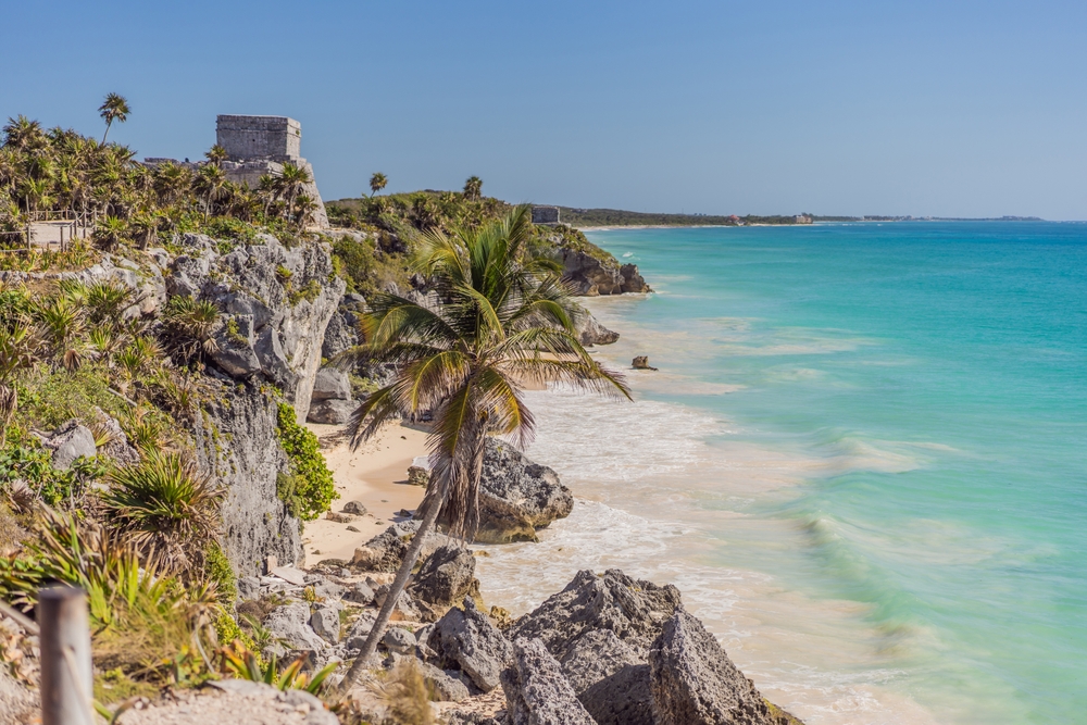 In Tulum, Mexico, the rocks of the terrain break into the waves on the shore, greenery poking through.