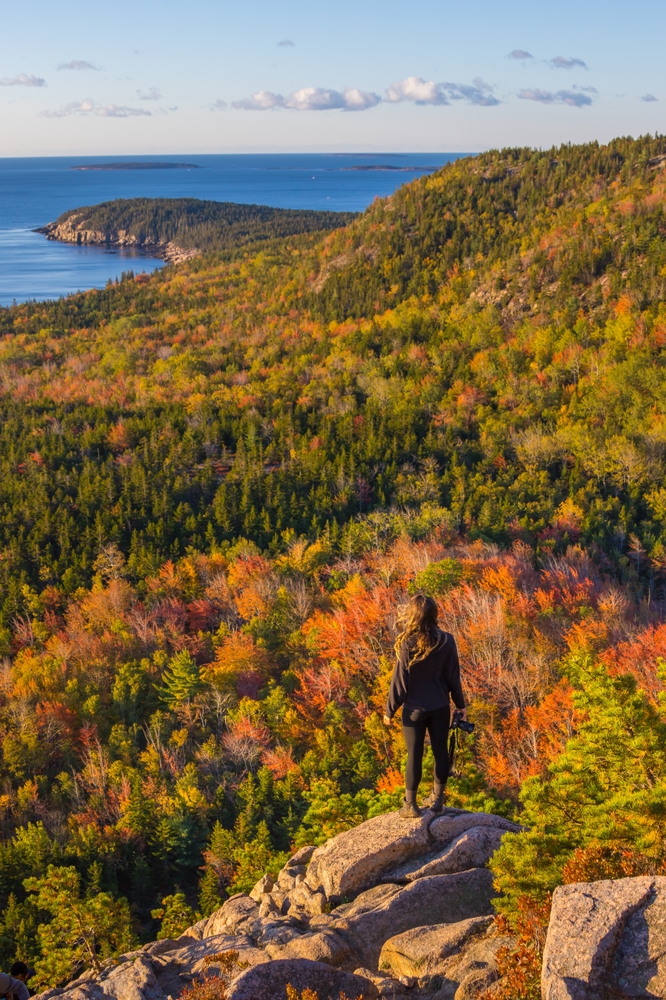 Female Hiker Black Shirt Stands on Rock Edge in Acadia National Park in fall. The article is about fall foliage weekend getaways.