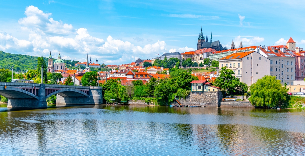 planning a trip to prague, photo of a river with a bridge on the left and historic buildings in the background