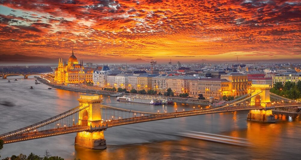 a sunset over the city of budapest, homes and government buildings are seen on the riverside, a long bridge reaches over the river.