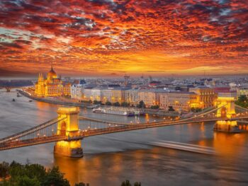 a sunset over the city of budapest, homes and government buildings are seen on the riverside, a long bridge reaches over the river.
