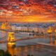 a sunset over the city of budapest, homes and government buildings are seen on the riverside, a long bridge reaches over the river.