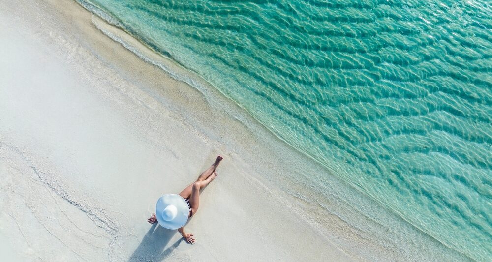 Woman from above wearing a white hat on the background of the sea and sunny beach. The article is about things to buy from Amazon before your beach vacation.
