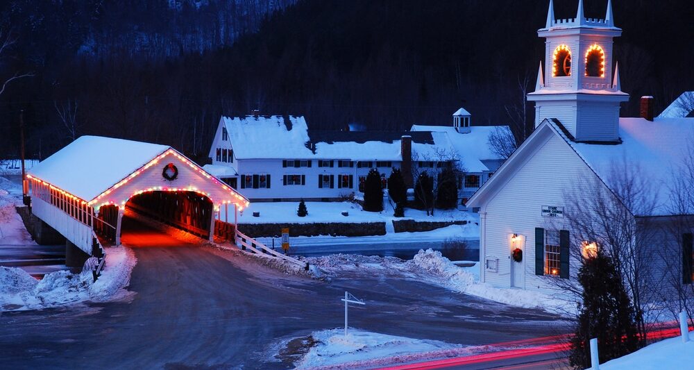 Snow covers a small town in New England in winter as the Christmas lights and decorations are illuminated on the church and covered bridge. The article is about towns in New England that feel like a hallmark movie.