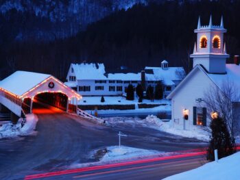 Snow covers a small town in New England in winter as the Christmas lights and decorations are illuminated on the church and covered bridge. The article is about towns in New England that feel like a hallmark movie.