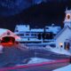 Snow covers a small town in New England in winter as the Christmas lights and decorations are illuminated on the church and covered bridge. The article is about towns in New England that feel like a hallmark movie.