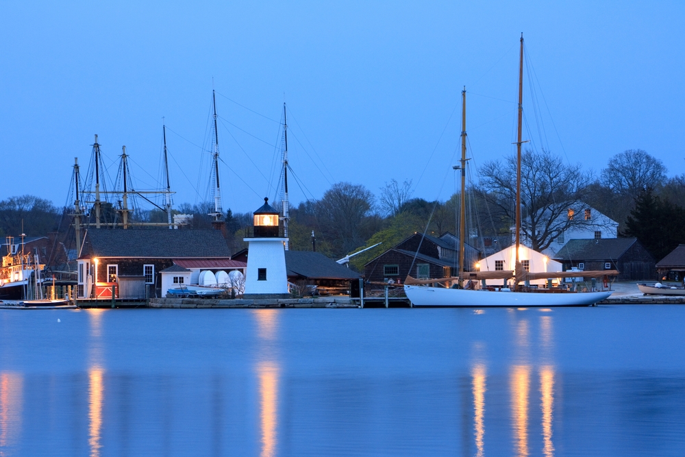 Scenic view of Mystic Seaport at night with a small white lighthouse and ships with tall masts. The article is about towns in New England that feel like a hallmark movie.