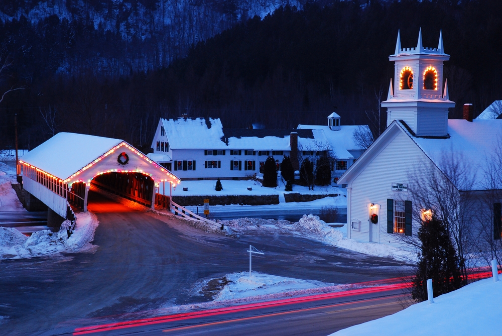 Snow covers a small town in New England in winter as the Christmas lights and decorations are illuminated on the church and covered bridge. The article is about towns in New England that feel like a hallmark movie.
