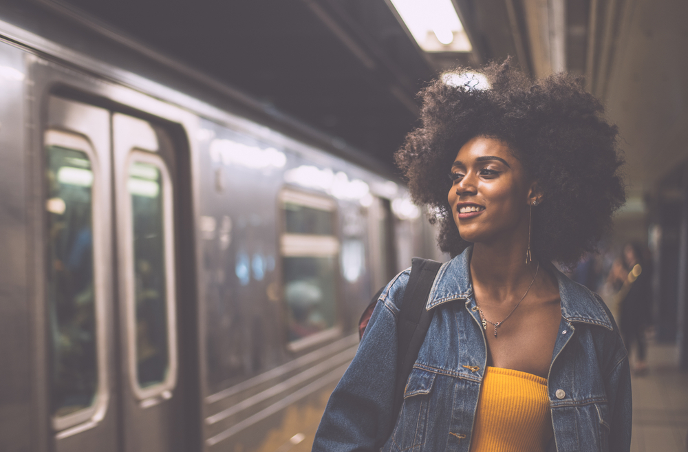 An African American woman in a yellow shirt and denim jacket smiles at the subway as it passes by her.