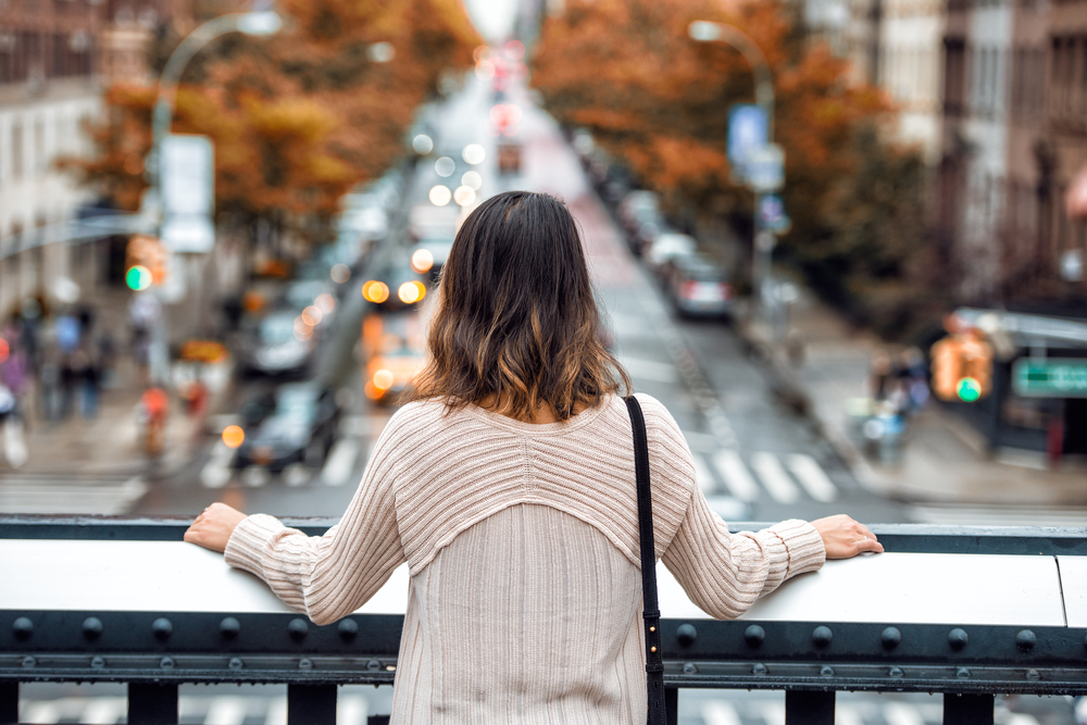 A woman in a wool sweater faces a street from a vantage point with her clutch over her shoulder as she enjoys the city.
