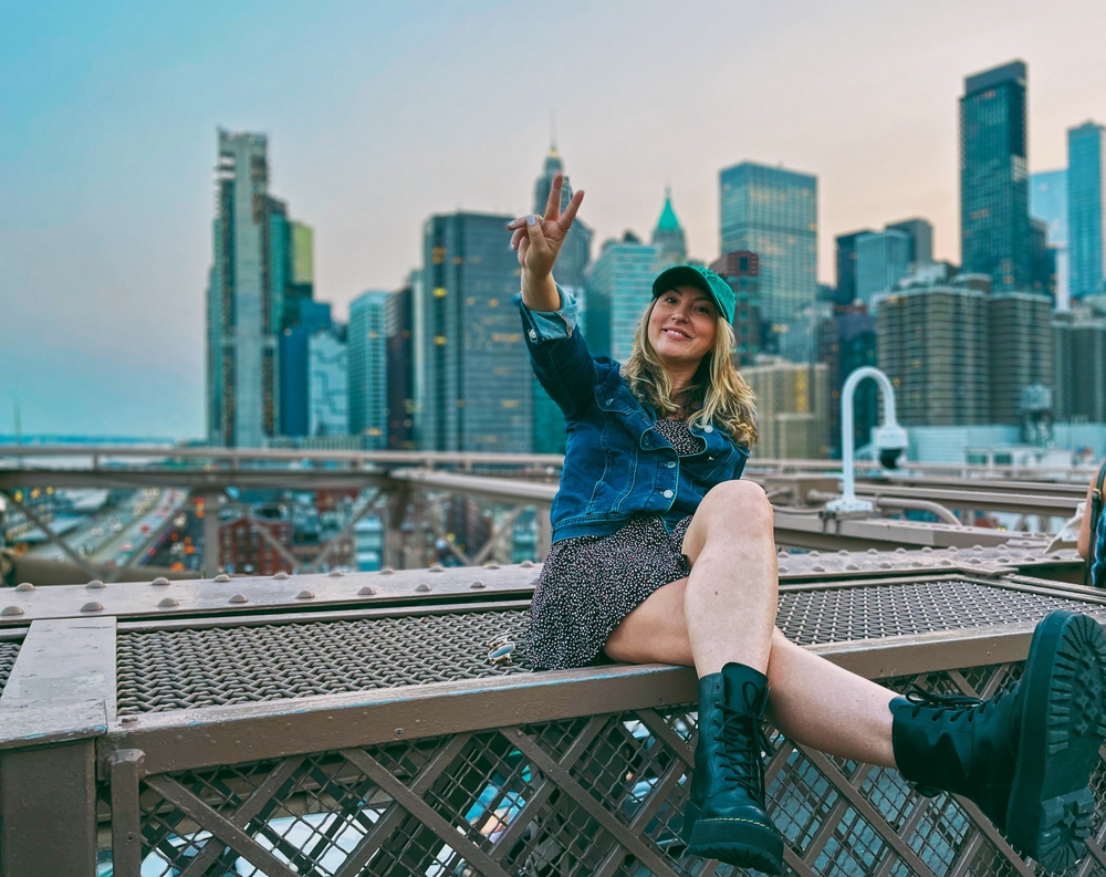 A woman in a midi dress, ball cap, and jean jacket throws a peace sign, knowing what to wear in New York City in September, with the city as a back drop behind her.