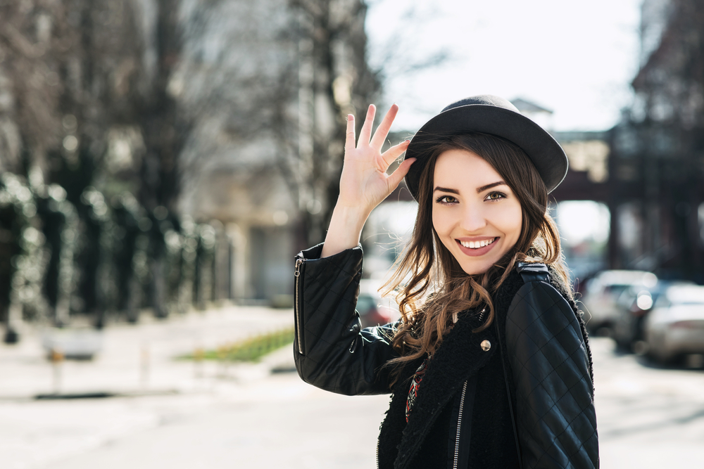 A woman smiles at the camera while tipping her hat and wearing a leather jacket, knowing what to wear in New York City in September.