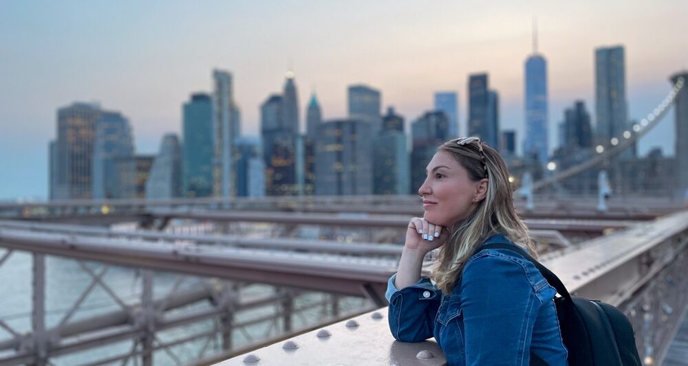 A woman overlooks the water on the Brooklyn bridge with a backpack on while wearing a denim jacket: she is well prepared to enjoy the city as she knew what to wear in New York City in September.