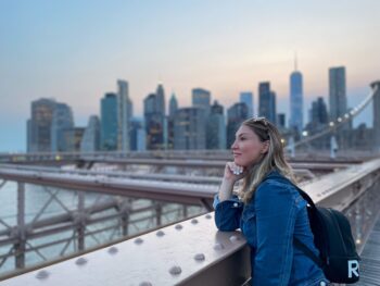 A woman overlooks the water on the Brooklyn bridge with a backpack on while wearing a denim jacket: she is well prepared to enjoy the city as she knew what to wear in New York City in September.