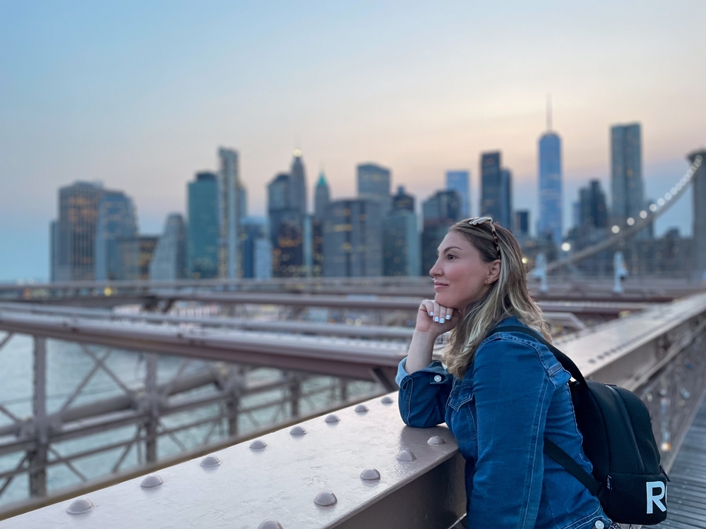 A woman overlooks the water on the Brooklyn bridge with a backpack on while wearing a denim jacket.