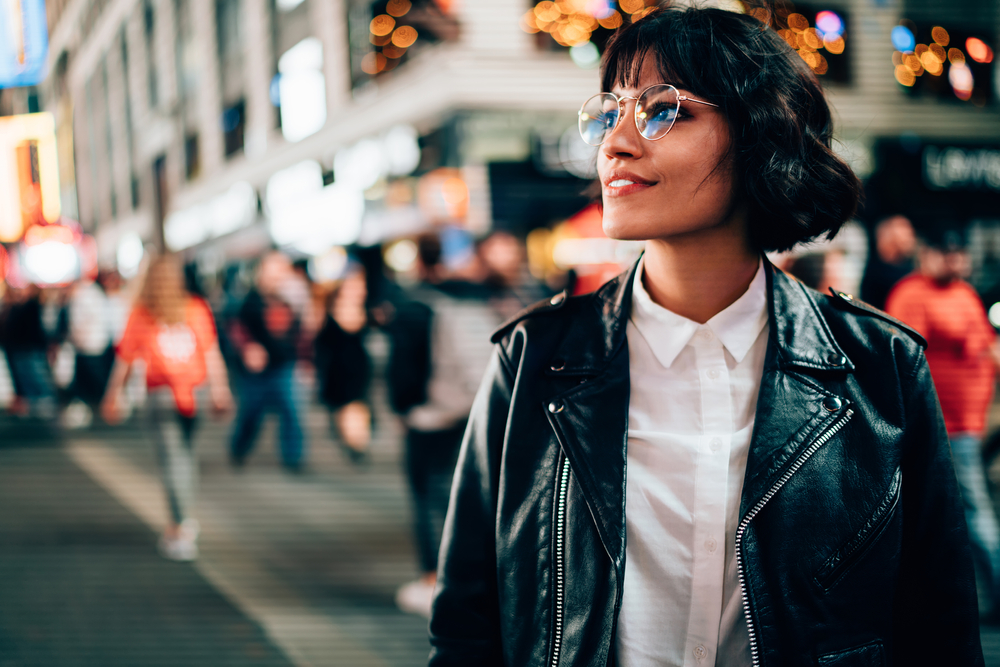 A girl channels her inner hipster and artsy style with round wire glasses, a button up blouse and a leather jacket as she walks through a busy intersection in NYC.