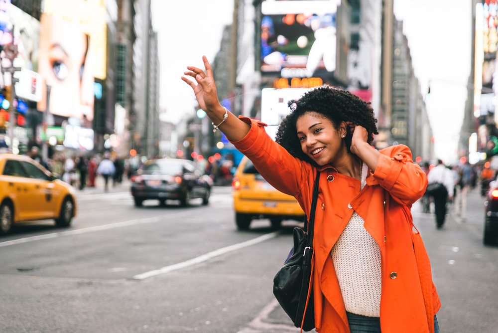 An African American woman waves down a taxi in the middle of the street in a knit sweater and orange jacket: she knew what to wear in New York City in September.