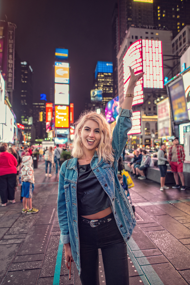 A woman in a jean jacket throws up her hand in the middle of time square because she knew what to wear in New York City in September.