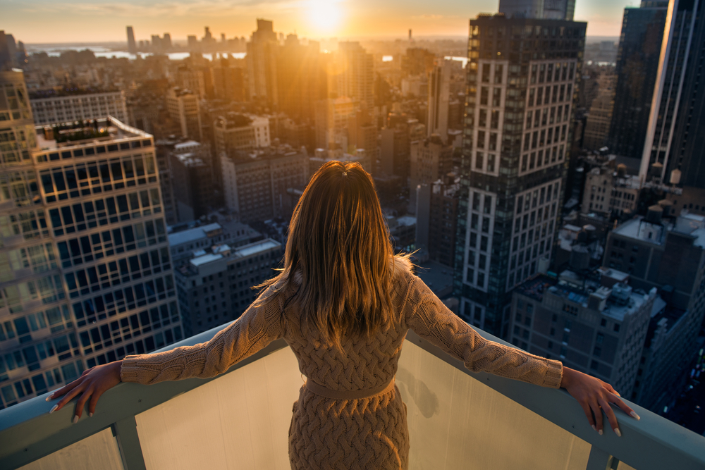 From behind, a woman stands on the corner of a rooftop bar in a sweater dress, arms spread as she looks down at the city, well prepared with what to wear in New York City in September.