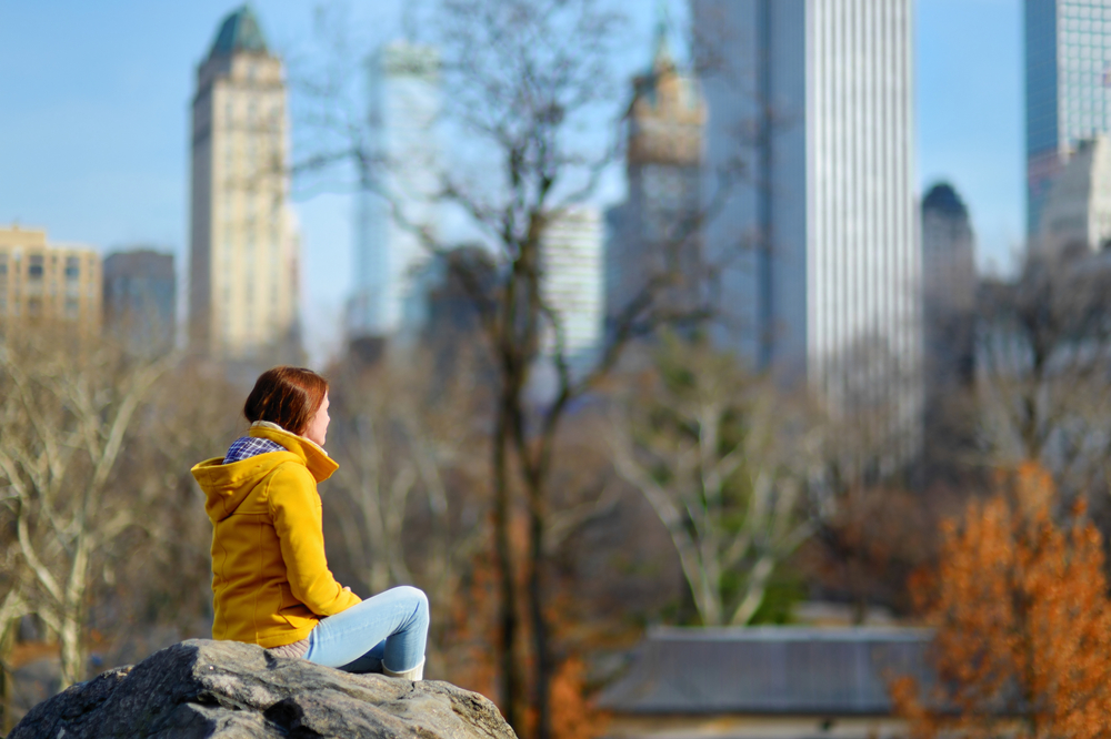 A woman sits on the edge of a rock in a yellow jacket and jeans as the trees around her turn yellow and gold.