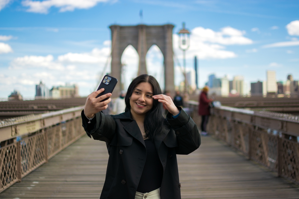 A woman with dark hair smiles for a selfie on a bridge with her dark blazer and black shirt underneath it, prepared and knowing what to wear in New York City in September.