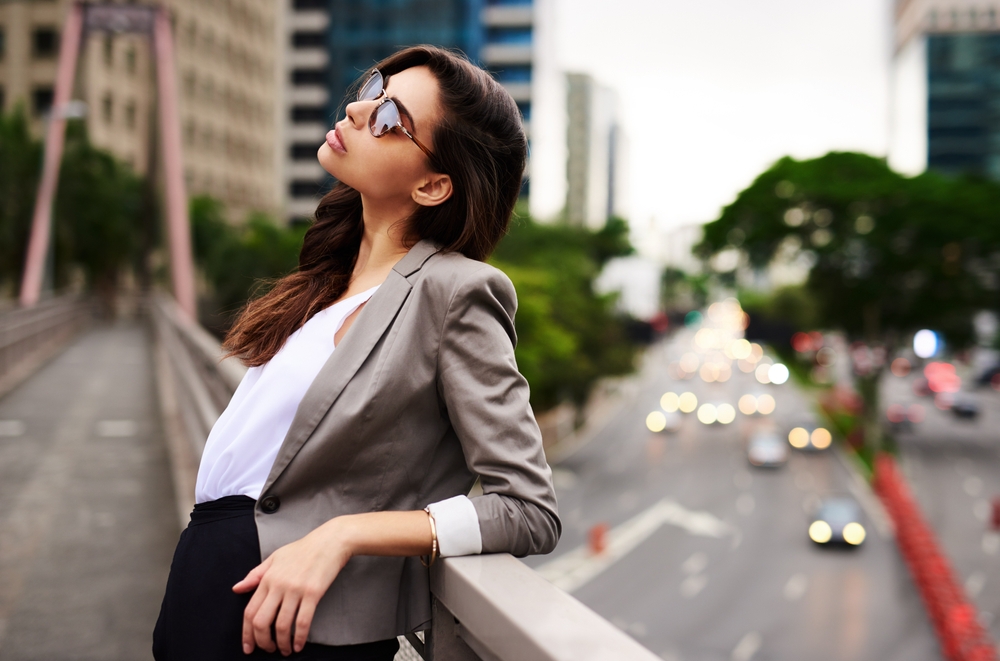 A woman leans against the edge of a bridge, comfortable in knowing what to wear in New York City in September in her blazer, silk blouse and sunglasses.