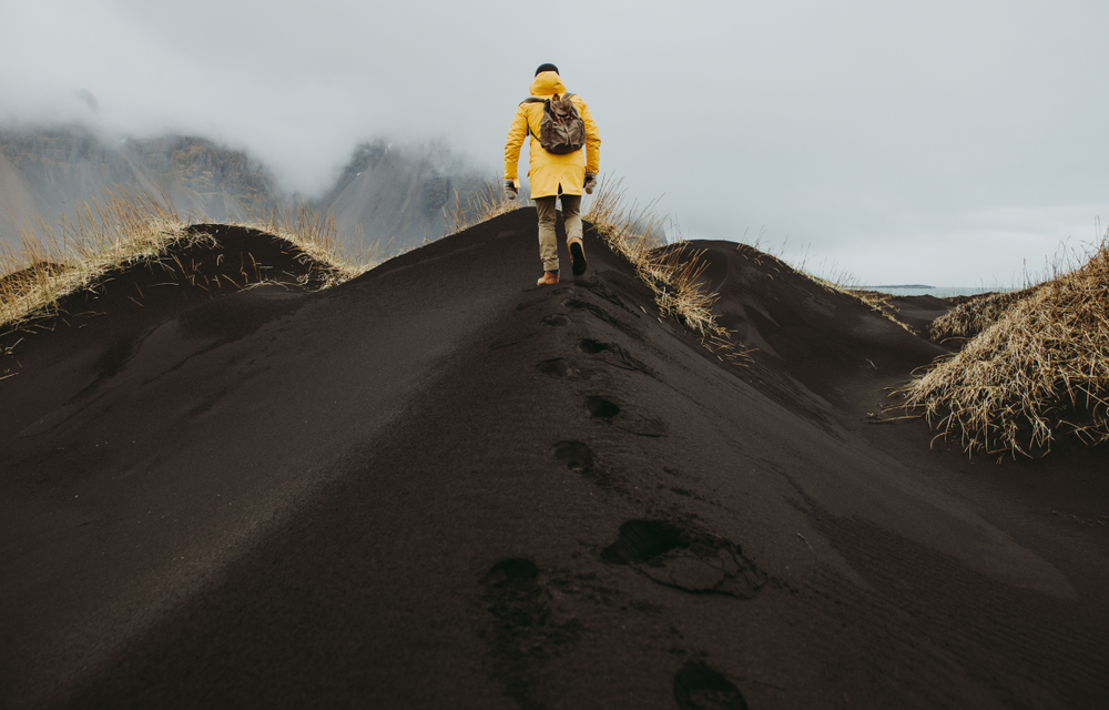 A tourist in a yellow parka and hiking pants climbs the black sand beaches in the fall, fog rolling beyond them.