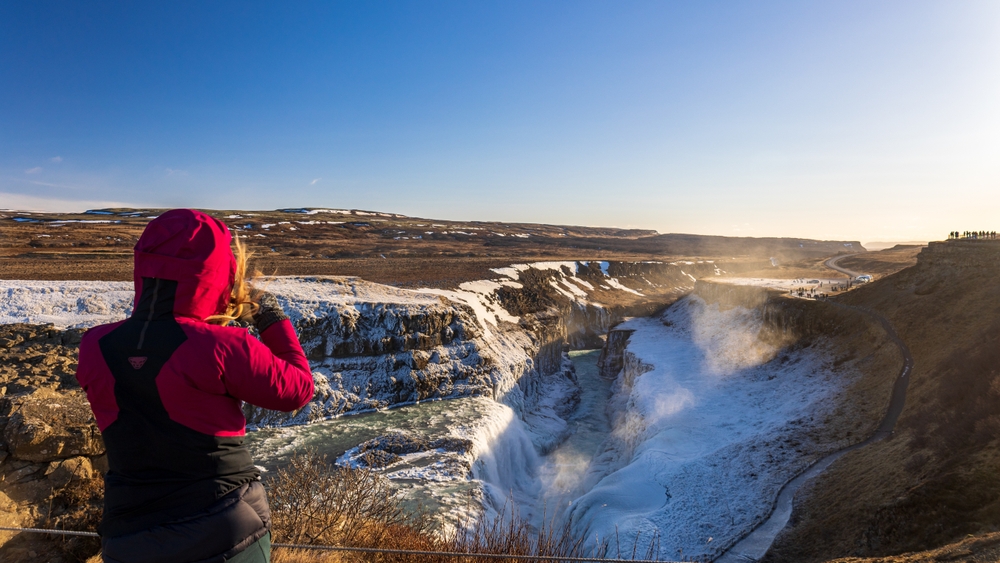 A woman takes a photo, overlooking a dip in the landscape that is full of waterfalls and snow, while wearing a pink windbreaker and gloves: she knew what to wear to Iceland in the fall.