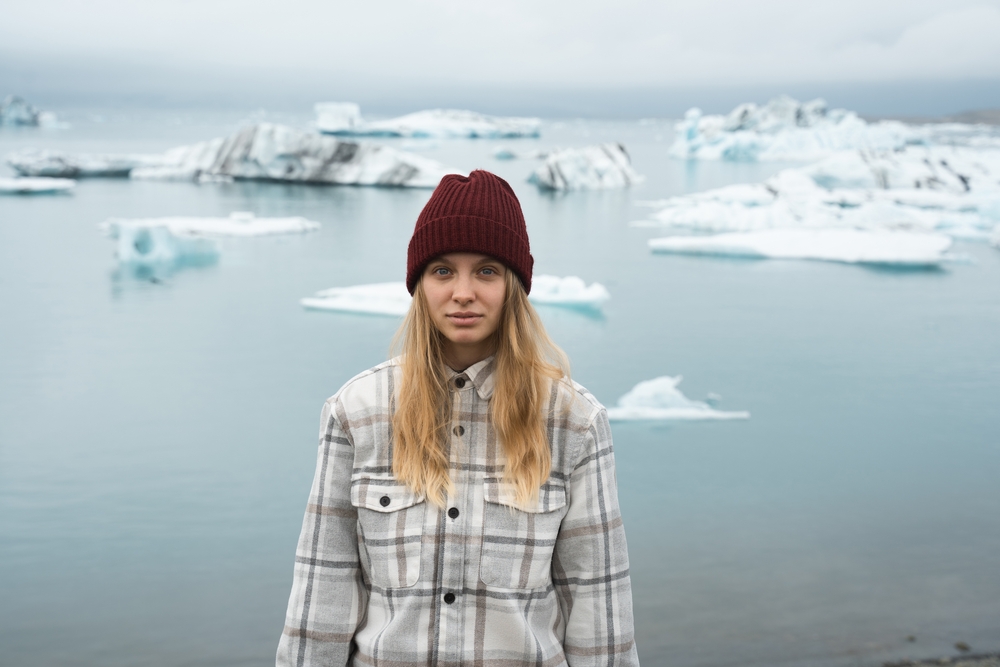 A blonde woman smiles at the camera as small chunks of ice pass beyond her: she wears a plaid flannel and beanie, perfect for what to wear to Iceland in the fall.
