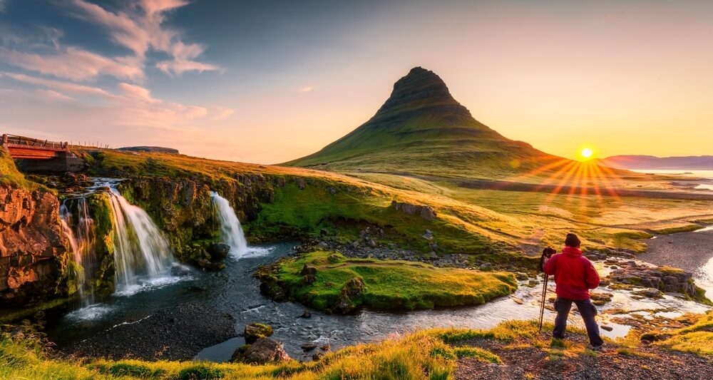 person wearing a red jacket and black pants stands in front of Kirkjufell mountain in iceland and there is a waterfall to the left and sunset to the right
